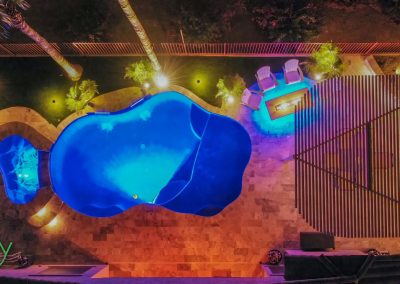 Aerial view of a brightly lit swimming pool surrounded by gardens and patio seating at night.