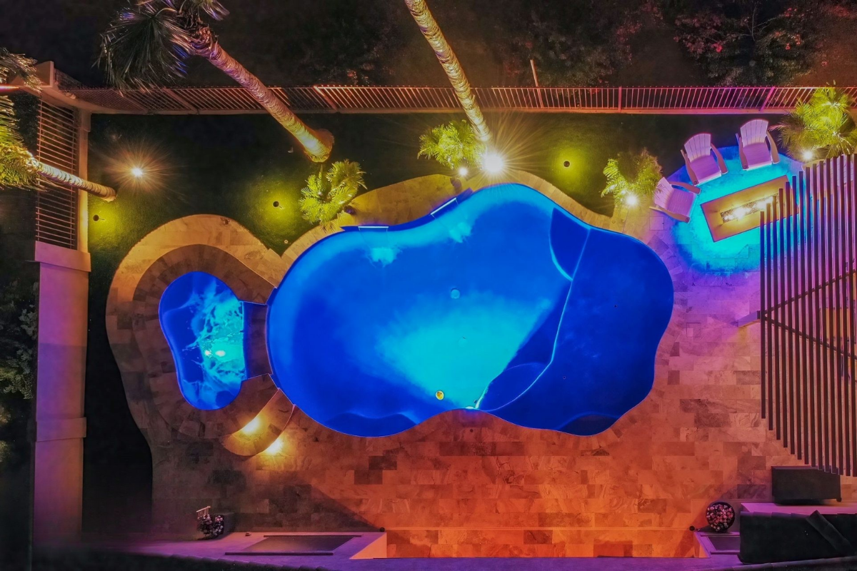 Aerial view of a brightly lit swimming pool surrounded by gardens and patio seating at night.